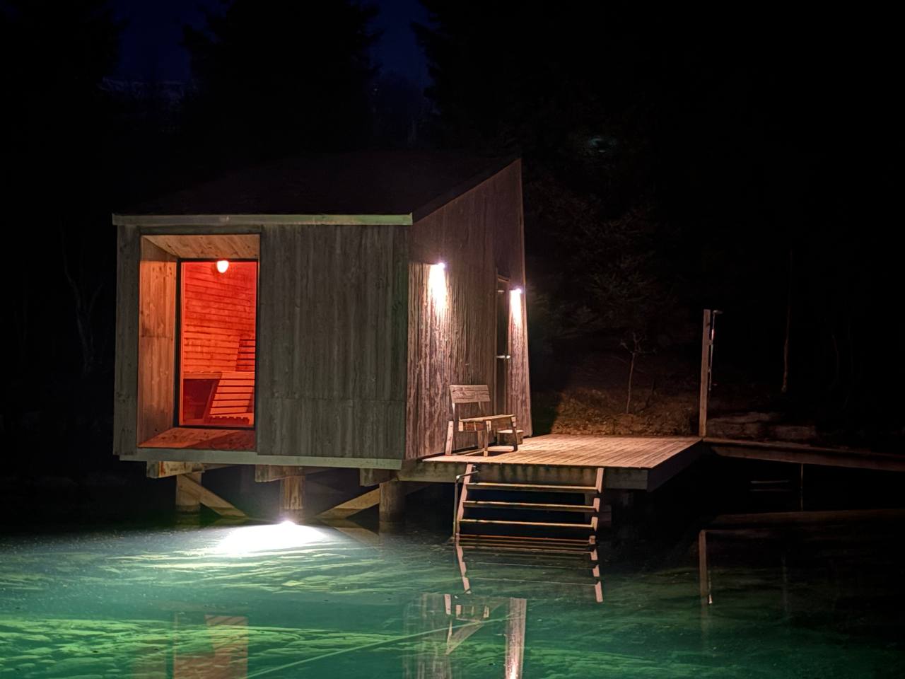 Sauna nad mořem v noci, dřevěná chata na pilotech, oknem svítí oranžovo-červeně, pod chatou tyrkysové moře a schůdky do vody