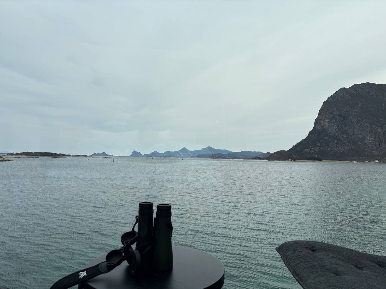 Šedý pohled oknem na fjord a hory, dalekohled na stolku