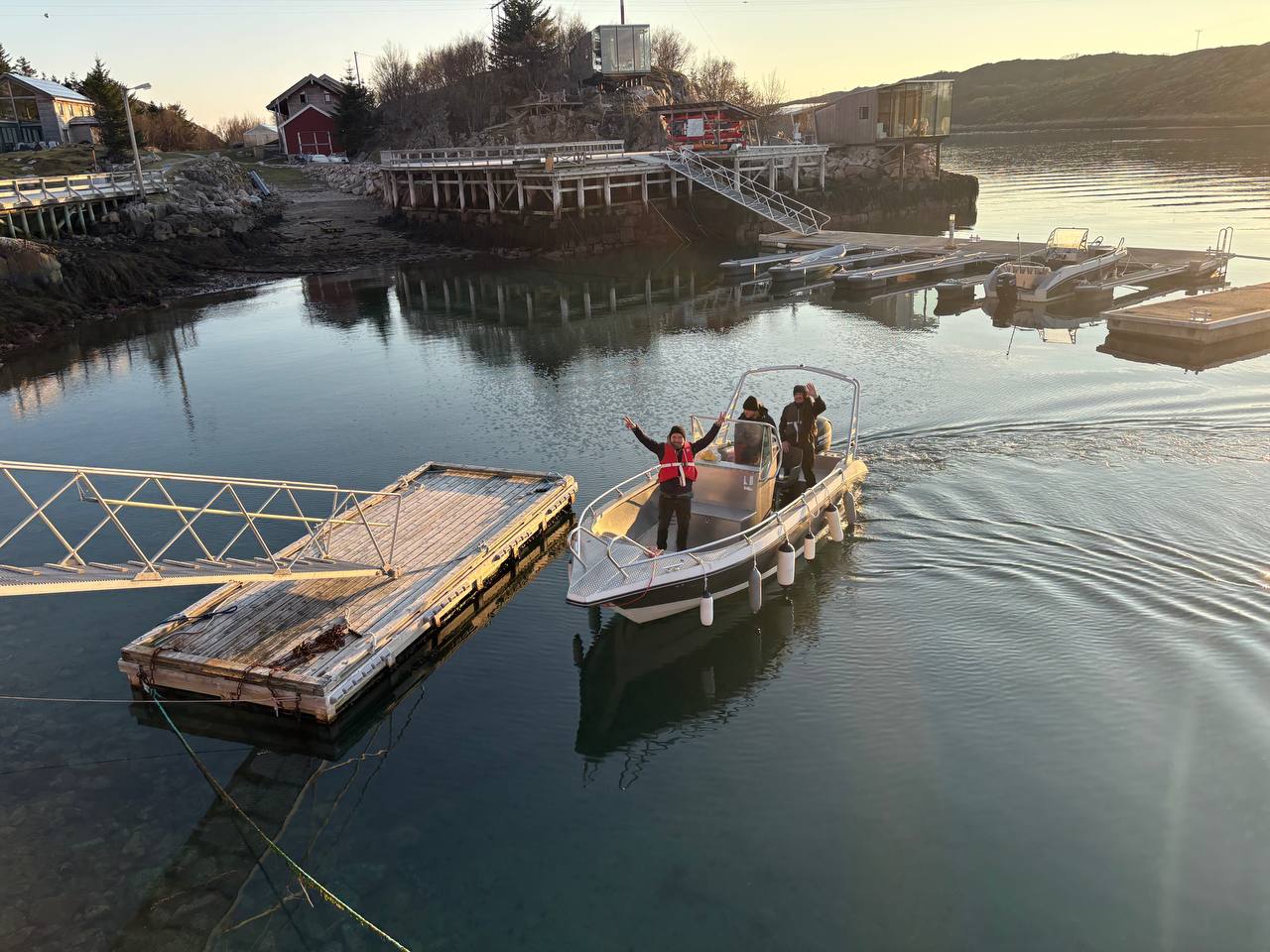 Dronový pohled shora na hliníkový shuttle u dřevěného mola Manshausen, dva lidé na palubě, jeden mává nahoru, červená bouda na břehu, zlatá hodina nad fjordem