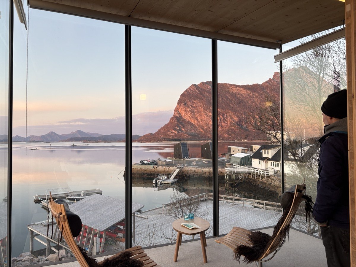 Postava u panoramatického okna kabiny, zlaté večerní světlo, výhled na fjord a zasněžené hory