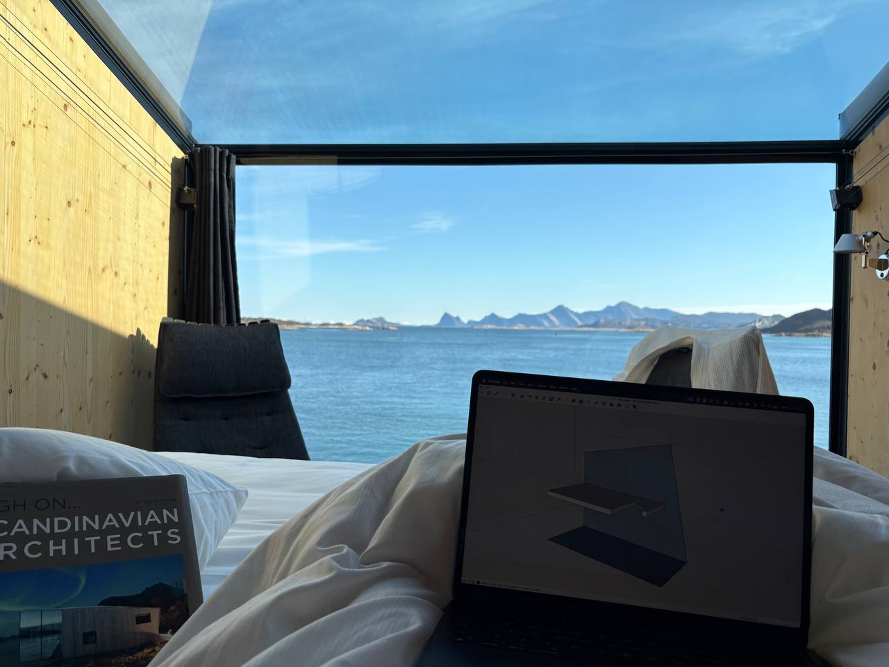 Pohled z postele v ohořelé boudě, na bílé peřině otevřený laptop s 3D modelem domu, vedle kniha Scandinavian Architects, za panoramatickým oknem fjord a hory v dopoledním slunci