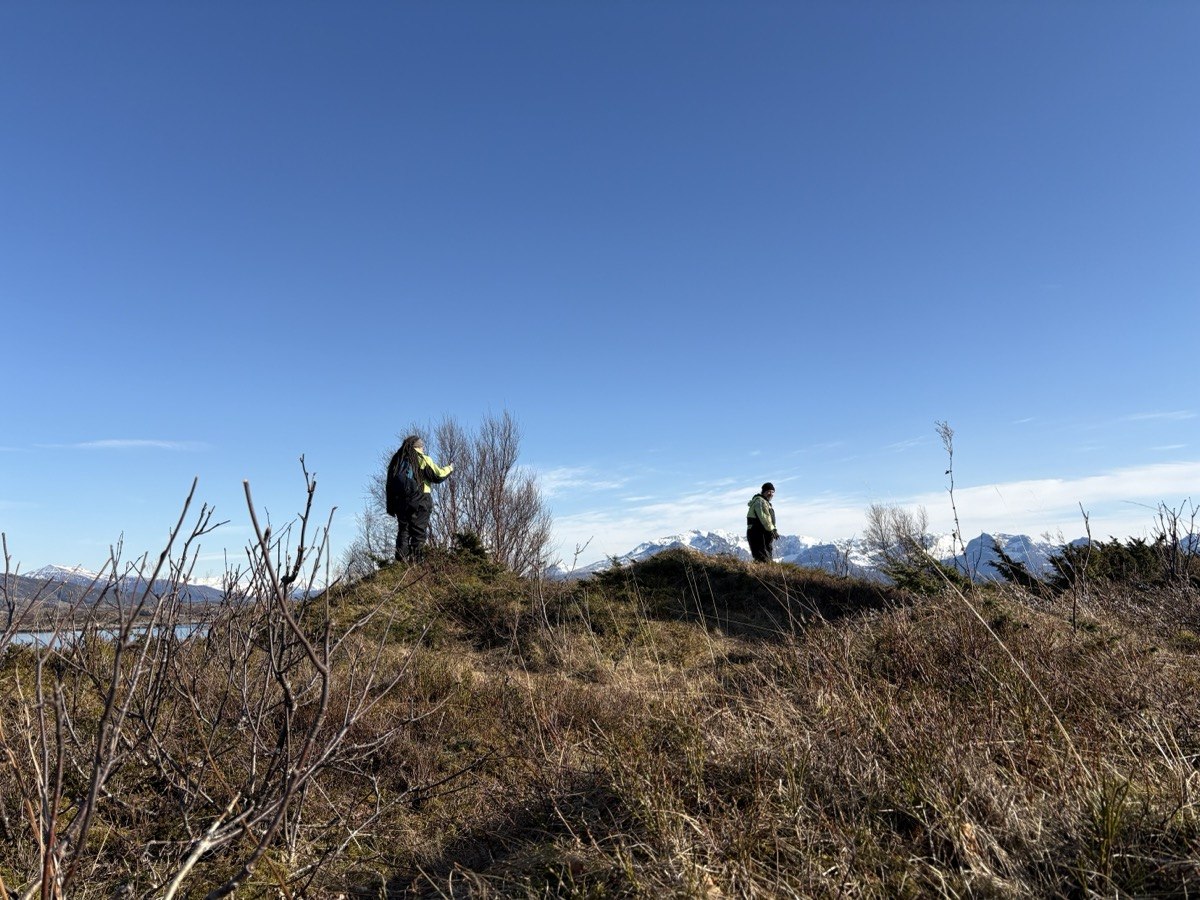 Dva lidé na kopci ostrova Måløya, suché trávy, zasněžené hory v pozadí přes fjord