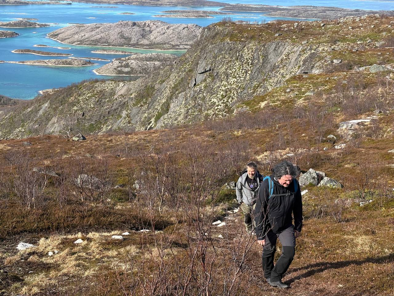 Dva lidé stoupají po skalnatém svahu, v pozadí souostroví a moře