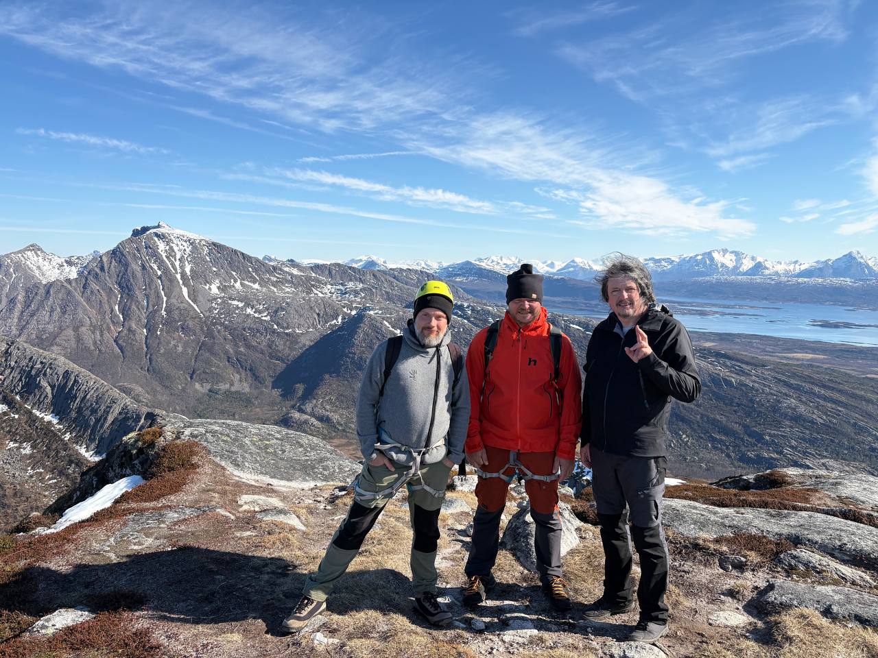 Tři lidé na vrcholu Sørskottinden — žlutá helma, červená bunda, tmavá bunda — s panoramatem souostroví a hor za zády