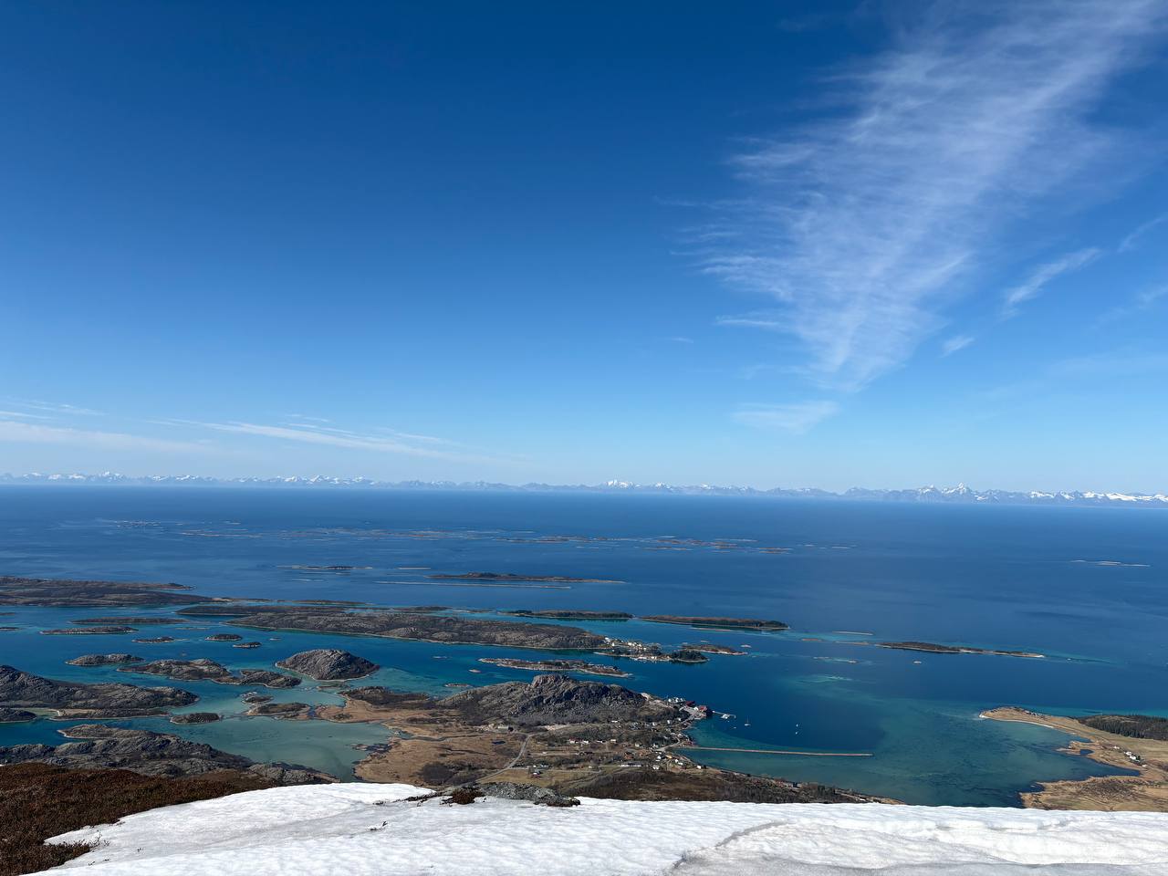 Výhled z vrcholu přes sněhové pole na ostrůvky v tyrkysovém moři, zasněžené hory na obzoru