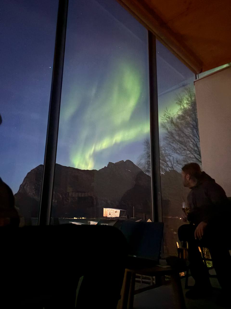Polární záře nad Manshausenem, zelený pás na obloze, silueta člověka s telefonem u okna, fjord a osvětlené domy