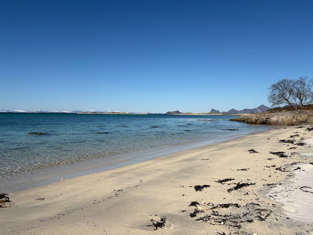 Bílá pláž s tyrkysovou vodou, skalnaté ostrůvky v pozadí, zasněžené hory na obzoru