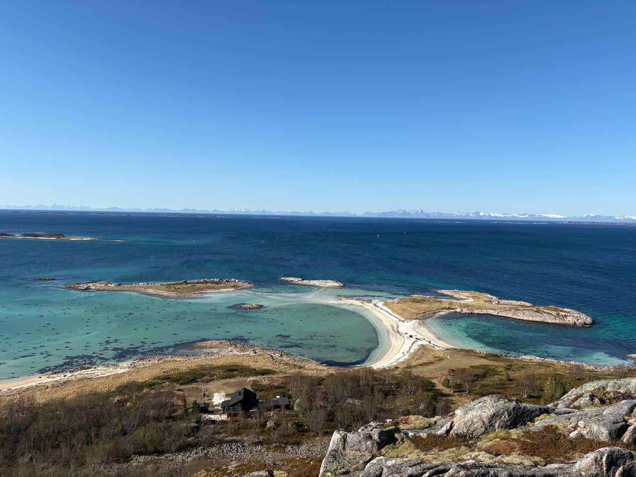 Tyrkysová zátoka s písečným přesypem spojujícím skalnaté ostrůvky, zasněžené hory v dálce