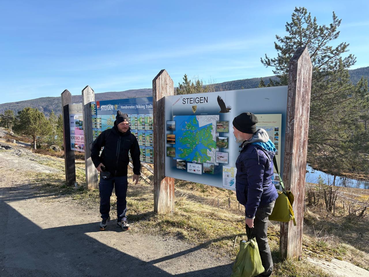 Dva lidé u informační tabule Steigen s výhledem na kopce a vodu
