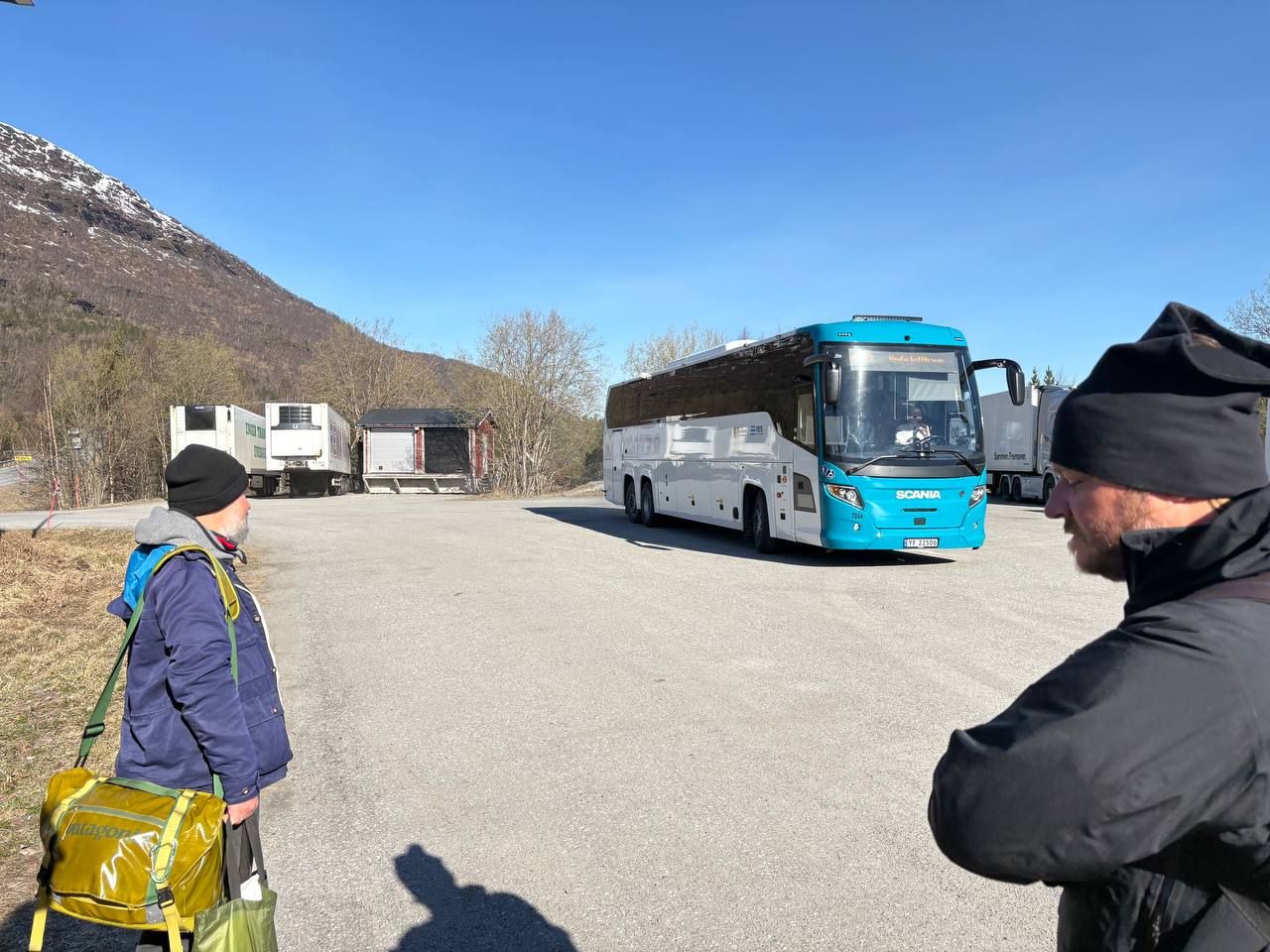 Lidé s batohy u autobusu na zastávce, zasněžené kopce v pozadí